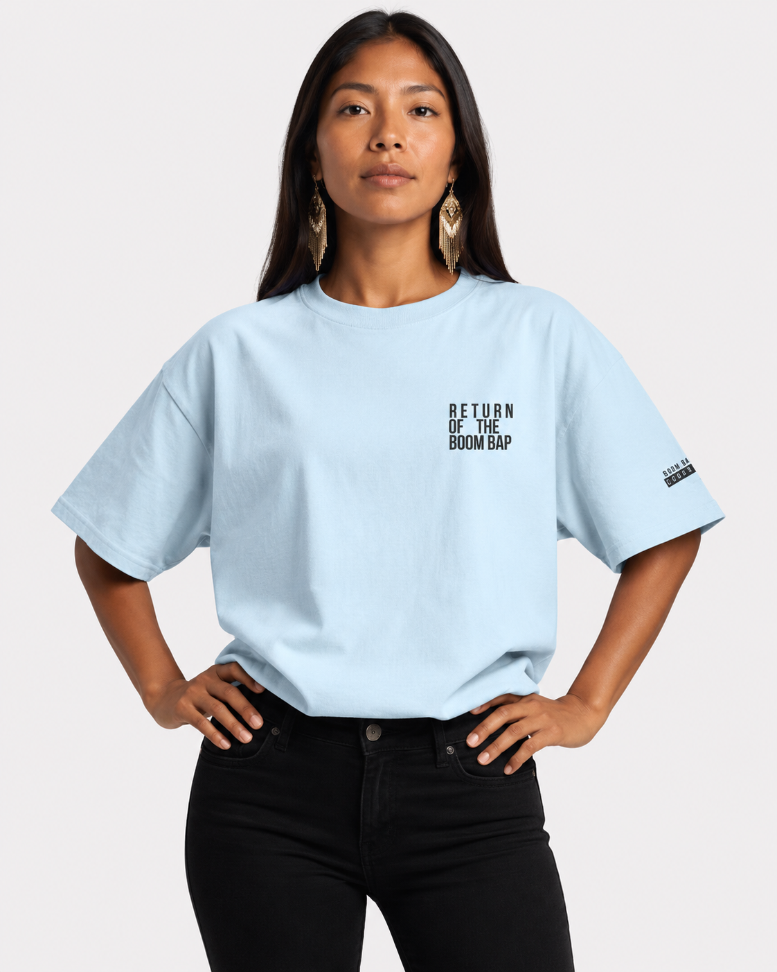 Front Lt. Blue A woman modeling a light blue oversized t-shirt with black text on the left chest reading 'RETURN OF THE BOM BAP' and a small black graphic on the sleeve, paired with black jeans against a light gray background.